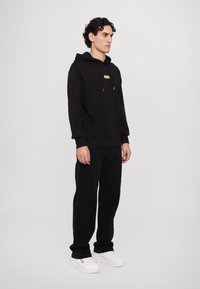 Black hoodie with a front gold logo patch, featuring a drawstring hood. Paired with black pants and white sneakers, standing against a neutral background.