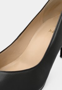 Svart läder högklackad sko med en slät textur, rund tå och subtilt inredning i beige. Har en kort klack.