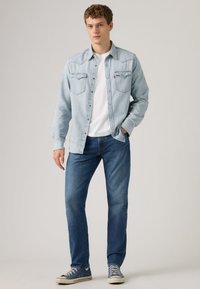 Light blue denim shirt with button-up design, two chest pockets, paired with dark blue jeans and gray sneakers. White t-shirt underneath.