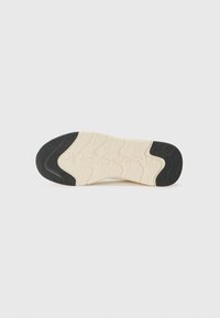 Semelle d'une chaussure avec une base en caoutchouc crème texturé et des zones de traction en caoutchouc noir, présentant un motif ondulé et un logo de la marque embossé.