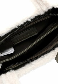 Taske med en beige faux fur yderside, uaf zipper for at afsløre et mørkt interiør med et lædermærke. Har sorte lynlåse og olivengrønne detaljer.
