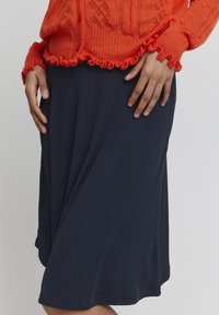 Meriharmaa ribbivartinen polvipituinen hame sileällä pintakuvioinnilla, jossa on levenevä muotoilu ja hienovarainen helmapitsi, yhdistettynä oranssiin teksturoituun paitaan.