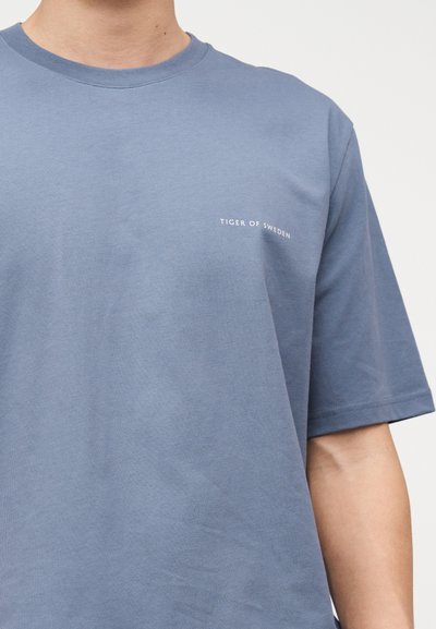 Μπλε κοντομάνικο T-shirt με μικρό λευκό κείμενο "Tiger of Sweden" στο αριστερό στήθος, που φοριέται από ένα άτομο σε απλό φόντο.