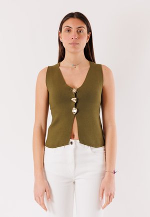 Donna con capelli lunghi e scuri che indossa un top senza maniche verde oliva con tre bottoni decorativi dorati, abbinato a pantaloni bianchi, in piedi contro uno sfondo semplice.