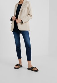 Blazer beige, top marino, jeans de mezclilla oscuros y sandalias negras con detalles trenzados. Materiales suaves, corte ajustado y jeans recortados.