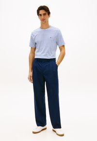 Ριγέ μπλε και λευκό t-shirt συνδυασμένο με σκούρο μπλε πλισέ παντελόνι. Το μοντέλο φοράει λευκά αθλητικά παπούτσια. Απλός σχεδιασμός, λεία υφάσματα.