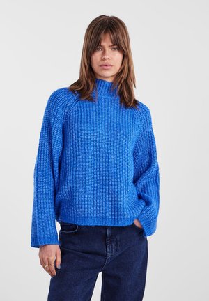Blue knit sweater with a high collar, textured weave, and wide sleeves, paired with dark jeans. The fabric appears soft and cozy.