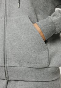 Szara hoodie na zamek z miękkiego materiału, z ribbowanymi mankietami i dolnym wykończeniem, bocznymi kieszeniami oraz subtelnym logiem w pobliżu kieszeni.
