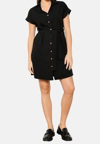 Czarna sukienka koszulowa z krótkim rękawem, kołnierzykiem, zapięciem na guziki, paskiem w talii i lekko luźnym krojem. Noszona z błyszczącymi czarnymi butami.