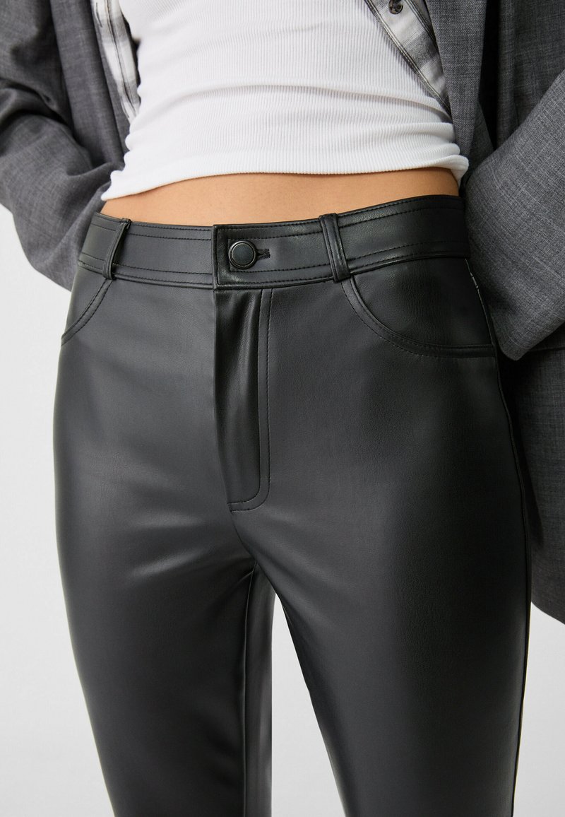 Pantalons en simili cuir noir avec une texture lisse, une coupe ajustée et des détails de couture visibles. Fermeture avec un bouton à l'avant et poches.