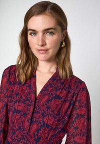 Abito a maniche lunghe in navy e bordeaux con motivo floreale. Presenta un profondo scollo a V e delle pieghe, realizzato in un tessuto leggero. Orecchini a cerchio dorati.