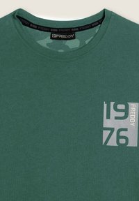 T-shirt in cotone verde con scollatura a girocollo, maniche corte e una grafica bianca con "1976" e "FREDDY" sul lato sinistro del petto. Texture morbida.