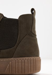 Olivgrön mocka sneakers med texturerad meshpanel, gummisula och vertikala ränder. Har en baktrycksklack och metallaccent.