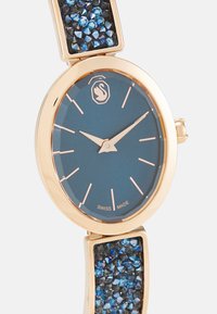 Reloj redondo con esfera azul, caja metálica de tono dorado y una correa que presenta cristales azules y negros como embellishment. Fabricado en Suiza.
