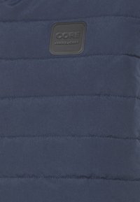 Marinblå vadderad väst med horisontell quiltad design. Har en texturerad svart fyrkantig etikett med texten "CORE". Slät tygfinish.