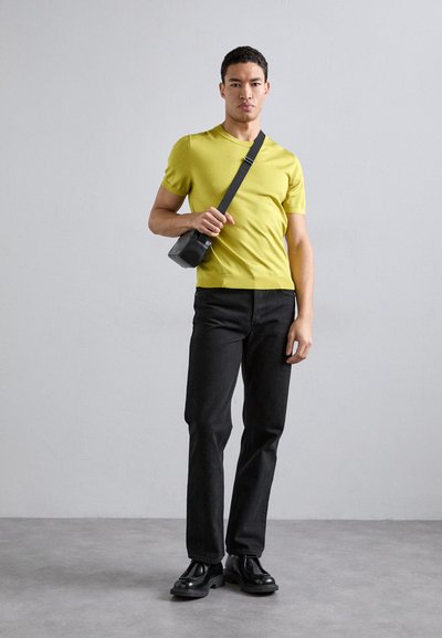 Tricou scurt de culoare verde lime, blugi negri cu croială dreaptă și botine negre. Geantă de umăr neagră cu curea. Fundal neutru.