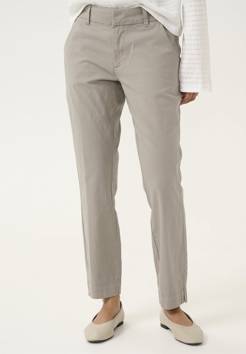 Femme portant un pantalon droit gris clair avec des poches latérales, assorti à des chaussures blanches enfilables et un haut blanc texturé à manches longues.