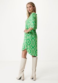 Grünes gemustertes Kleid mit kurzen Ärmeln, asymmetrischem Saum und lockerer Passform, kombiniert mit kniehohen beigen Stiefeln.