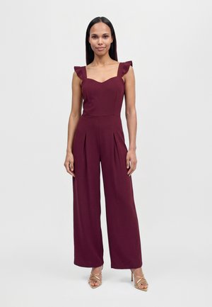 Une femme porte une combinaison bordeaux à jambes larges et à bretelles volantées, avec des talons dorés, sur un fond clair et uni.
