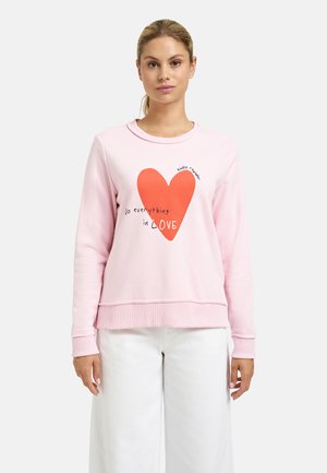 Young woman with blonde hair wears a light pink sweatshirt with a large red heart and text "do everything in LOVE" standing against a white background.