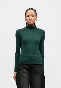 Πράσινο μακρυμάνικο τοπ με ζιβάγκο από απαλό ύφασμα· εφαρμοστή σχεδίαση με λεία υφή, σε συνδυασμό με σκούρο τζιν παντελόνι.