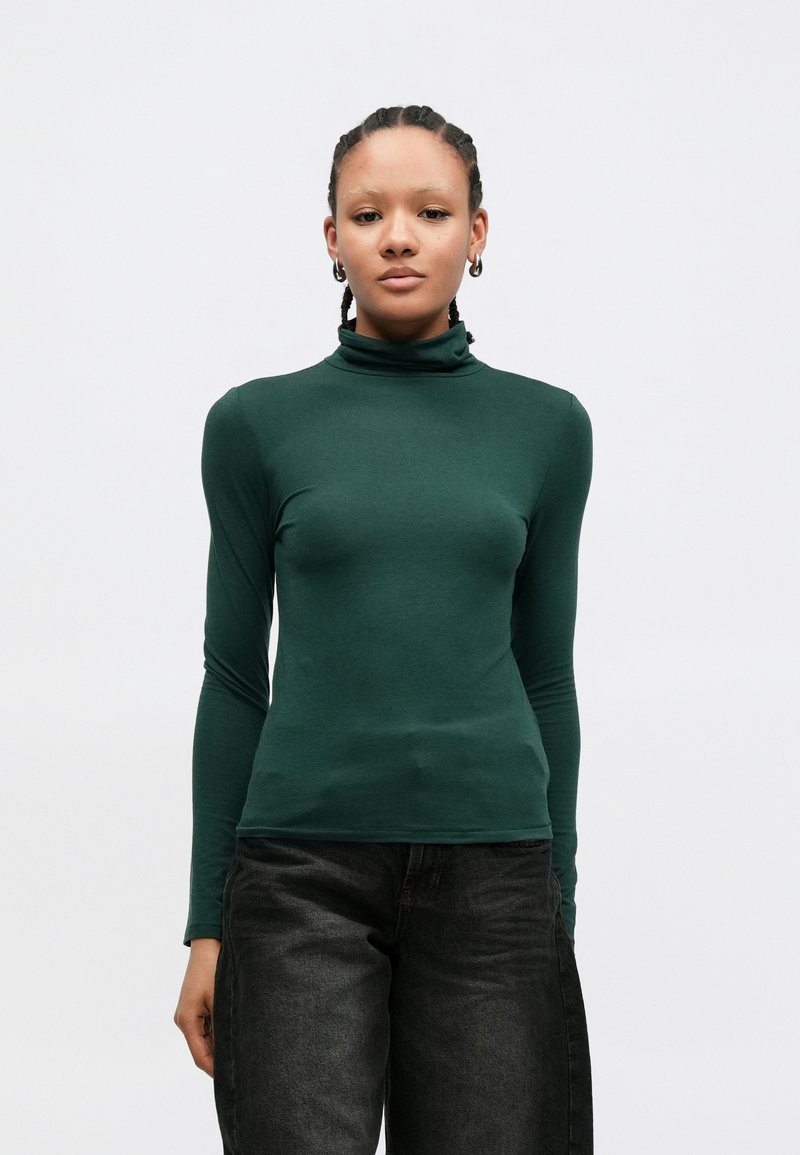 Πράσινο μακρυμάνικο τοπ με ζιβάγκο από απαλό ύφασμα· εφαρμοστή σχεδίαση με λεία υφή, σε συνδυασμό με σκούρο τζιν παντελόνι.