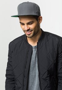 Veste bomber noire matelassée sur un t-shirt gris, accompagnée d'une casquette grise avec une visière verte. Design épuré sans logos visibles.