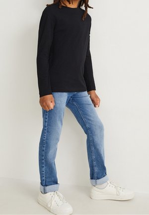 Henkilö, jolla on päällään musta pitkähihainen paita, vaaleansiniset käännetyt farkut, valkoiset sukat ja valkoiset tennarit, seisomassa yksiväristä taustaa vasten.