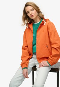 Giacca a vento arancione con cappuccio, polsini elasticizzati e tasca laterale con cerniera. Indossata sopra una felpa verde e jeans chiari.