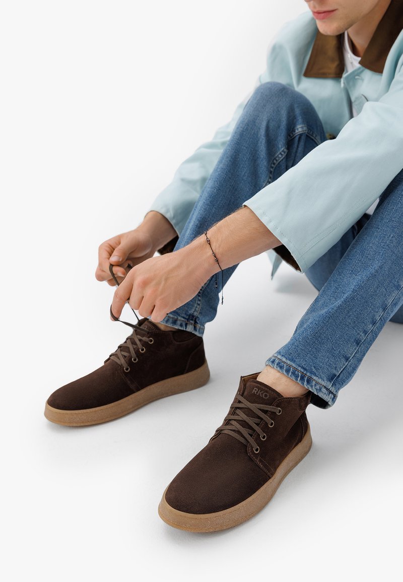 Stivaletti in suede marrone con suola in gomma beige e lacci piatti. Presentano dettagli cuciti e un piccolo logo sul lato esterno.