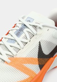 Fehér sportcipő texturált hálós felsőrészsel, narancssárga díszítéssel, fekete Nike logóval, lapos fehér fűzőkkel és színes cimkével a nyelvén.