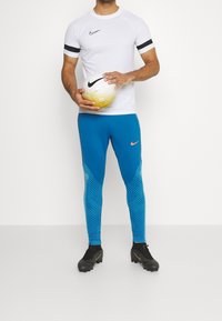 Nike sportoutfit bestaande uit een witte T-shirt met korte mouwen, felblauwe leggings met een gestructureerd patroon en zwarte voetbalschoenen, terwijl hij een voetbal vasthoudt.