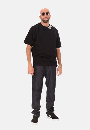 T-shirt oversize noir avec un détail de logo, pantalon en denim foncé et baskets noires. Les accessoires comprennent des lunettes de soleil rondes et une boucle d'oreille en argent.