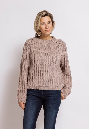 Woman with short blonde hair wearing a loose, chunky knit beige sweater and dark blue jeans standing against a plain light background.