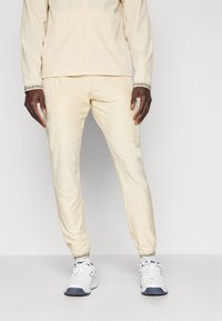 Beige treningsjakke og bukse-sett med tettsittende mansjetter, teksturert stoff og sidepaneler. Kombinert med hvite joggesko med blå detaljer.