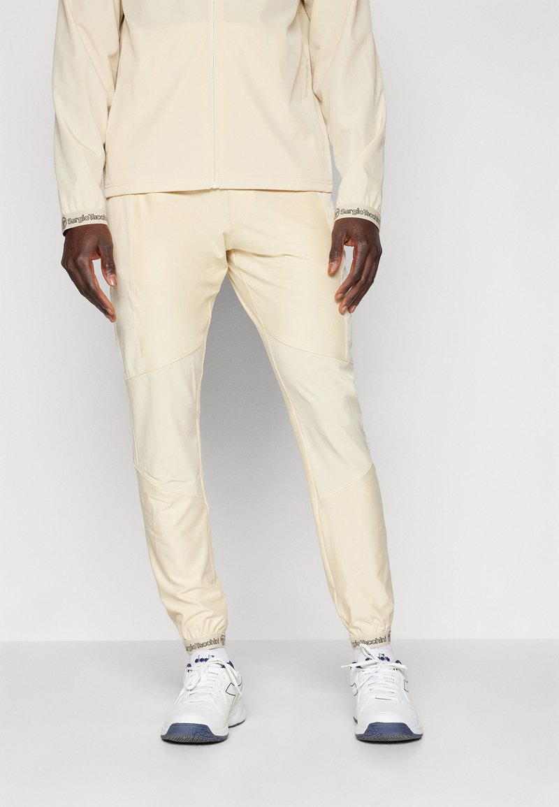 Beige treningsjakke og bukse-sett med tettsittende mansjetter, teksturert stoff og sidepaneler. Kombinert med hvite joggesko med blå detaljer.