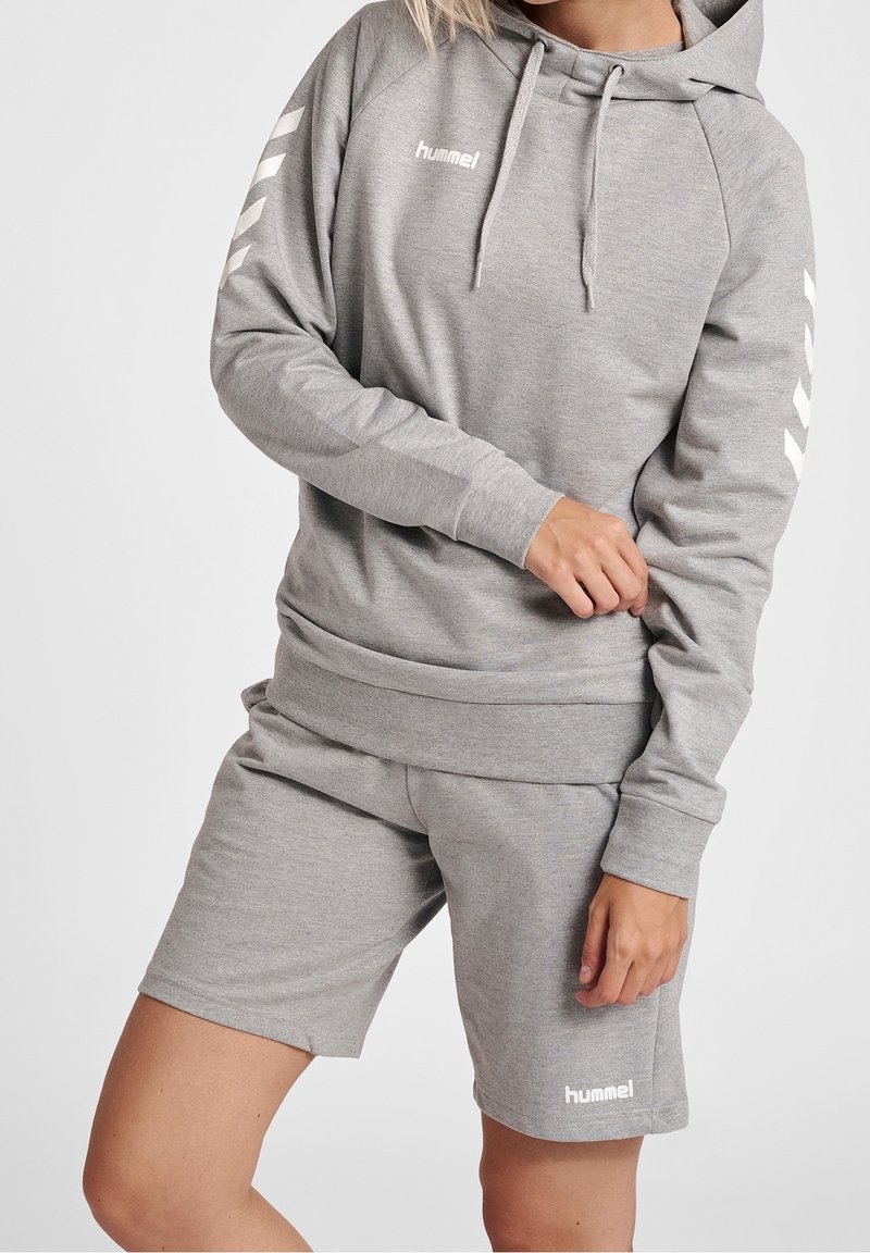 Personne portant un sweat à capuche hummel gris clair et un short assorti avec des motifs chevrons blancs sur les manches et des logos sur la poitrine et la jambe.