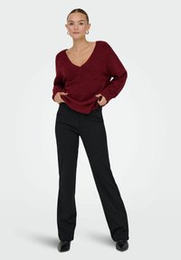 Pull en tricot bordeaux avec un décolleté en V profond et des poignets côtelés, associé à un pantalon noir à jambe droite et des chaussures noires à bout pointu.