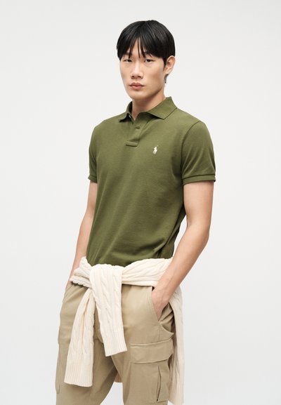 Νέος άνδρας που φοράει πουκάμισο πόλο ελιάς, μπεζ παντελόνι cargo και πλεκτό πουλόβερ κρεμ δεμένο γύρω από τη μέση, στέκεται με τα χέρια στις τσέπες.