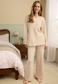 Beežist ribaga loungewear komplekt, mis koosneb pika varrukaga topsist ja laienevatest püksidest. Pehme tekstuur, mugav lõige. Näha on heledate toonidega sisustusega tuba.