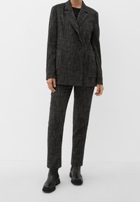 Blazer textured nero e grigio con design doppiopetto, abbinato a pantaloni su misura coordinati e stivali neri alla caviglia.