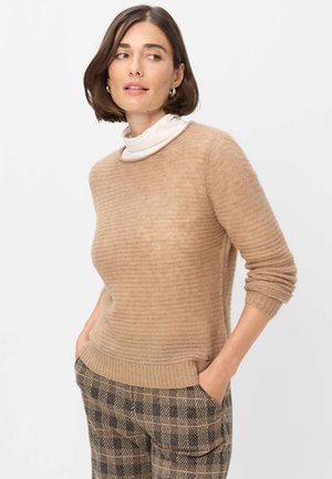 Kvinde med kort brunt hår iført en beige strikket sweater over en hvid højhalset trøje og ternede bukser, hænderne i lommerne, ser til siden.