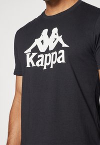 Černé tričko s bílým logem Kappa, které zobrazuje dvě sedící siluety zády k sobě a text "Kappa" pod nimi.