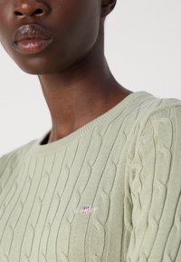 Pullover a maglia verde chiaro con motivo a trecce, colletto rotondo e polsini a coste. Presenta un piccolo logo ricamato sul petto.