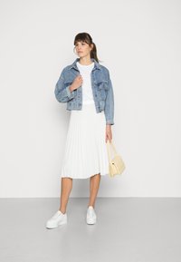 Giacca in denim, t-shirt bianca, gonna bianca plissettata, sneakers bianche e piccola borsa trapuntata beige, in piedi contro uno sfondo neutro.