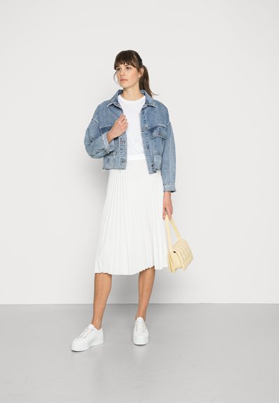 Veste en jean, t-shirt blanc, jupe plissée blanche, baskets blanches et petit sac beigne matelassé, se tenant devant un fond uni.