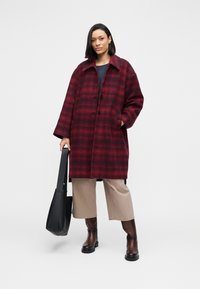 Red plaid wool coat with a collared neckline and button closures, paired with wide beige trousers and brown knee-high boots. Black tote bag.