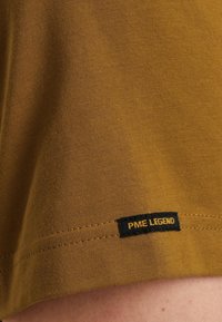 Tielko v horčicovej farbe vyrobené z bavlny, s hladkou textúrou a dokončenými okrajmi. Na ňom je výrazne umiestnená čierna nášivka s nápisom "PME LEGEND".