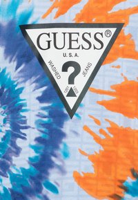 Logo triangulaire noir avec le texte "GUESS U.S.A." sur un tissu en tie-dye multicolore comportant des teintes de bleu, orange et bleu clair.