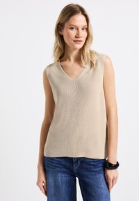 Top senza maniche beige in maglia con scollatura a V, caratterizzato da un design testurizzato e dettagli di cucitura verticale, abbinato a jeans in denim blu e un bracciale nero.